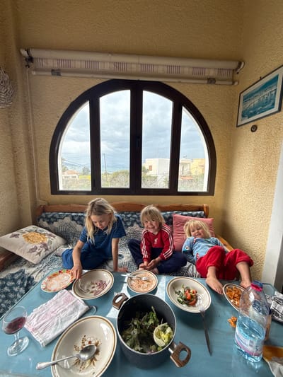Three young children are seated on a blue and white patterned daybed in a room with textured, light yellow walls, situated in Aegina, Greece. An arched, dark-framed window in the background reveals a cloudy sky, residential buildings, vegetation, and a distant body of water, with rain streaks visible on the glass. Above the window, a rolled-up striped blind is present. To the right of the window, a framed blue-toned coastal landscape print hangs, with a circular object below it.

On the left, a blonde child in a blue striped shirt and trousers sits cross-legged, looking down at an oval bowl on a light blue table, holding a fork. In the center, a blonde child in a red and white striped long-sleeved shirt sits cross-legged, smiling directly at the camera. On the right, a blonde child in a blue patterned top and red trousers reclines with a pink cushion, also smiling at the camera, their bare feet near a plate.

The light blue table in the foreground holds various items: a dark pot containing green salad (lettuce, red onion), multiple oval and round dishes with remnants of food (pasta, salad, sauces), several forks and spoons, a clear glass with red liquid, a crumpled white and pink striped cloth, and a large clear plastic water bottle. The scene suggests a meal has recently taken place or is ongoing.