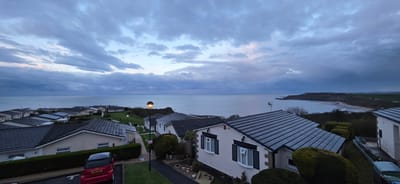 The image depicts a coastal residential area in Osgodby, United Kingdom, under a cloudy sky at dusk. The sea stretches to the horizon, with a headland visible in the distance. In the foreground, rows of similar single-story houses with gray tiled roofs are interspersed with landscaped lawns and pathways. A red compact car is parked near one of the houses. Street lights are illuminated, one with a spherical lamp prominently visible. The scene suggests a quiet, possibly resort-oriented, residential area near the coast.
