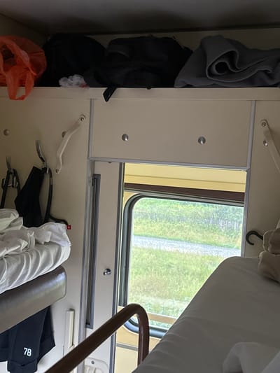 This image depicts the interior of a train compartment, likely in Shalya, Russia. The compartment contains two berths, one above the other. The upper berth is covered with white bedding, and a brown luggage rack is visible above it, holding several bags, including an orange plastic bag and two dark-colored backpacks. Below the upper berth, on the wall, are two metal hooks. A black garment, possibly pants, with the number "78" printed on it in white, hangs from a hook on the lower berth. Adjacent to the lower berth is a window looking out onto a landscape with green foliage and a gravel path. A metal handrail for the upper berth is visible in the foreground, and a portion of the lower berth is also visible. The walls and ceiling of the compartment are a light beige color. There are no people visible in the image.