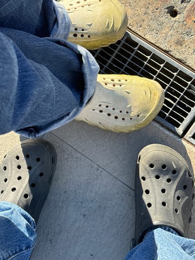 The image captures a ground-level view of four feet on a paved surface in Lipomo, Italy. Two individuals are represented by their lower extremities, clad in blue denim jeans and Crocs clogs. In the upper right portion of the frame, a pair of feet wearing light yellow, visibly soiled Crocs is positioned next to a rectangular metal drainage grate. Below and to the left, another pair of feet is visible, wearing dark grey Crocs. The ground consists of light grey square tiles with distinct grout lines. Strong, direct sunlight illuminates the scene, casting prominent shadows from the feet and clothing. The scene depicts a static moment with no overt action or interaction between the subjects.