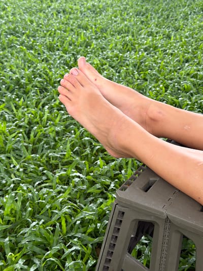 The image shows a close-up view of a person's bare feet and lower legs resting on a dark green folding stool. The background is a dense expanse of lush green grass, indicating an outdoor setting. The skin on the legs shows some light scarring. The lighting suggests it is daytime. The location is identified as Tambon San Phi Suea, Thailand.