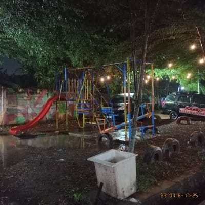 A playground, illuminated by string lights, stands in a yard at night. A slide with a red ramp sits near a climbing structure made of colorful metal bars. Several old tires are scattered around the playground area. A large, weathered, white planter box rests beside the play area, which appears damp, suggesting recent rain. In the background, a vehicle with visible lettering can be seen. The image is dark and somewhat blurry, giving a nighttime atmosphere.