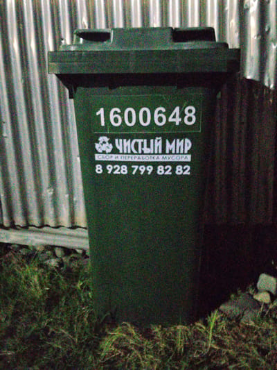 In Plievo, Russia, a dark green garbage bin stands against a corrugated metal fence. White lettering on the bin reads "1600648", a recycling symbol, "ЧИСТЫЙ МИР" (meaning "Clean World"), "СБОР И ПЕРЕРАБОТКА МУСОРА" (meaning "Waste Collection and Recycling"), and the phone number "8 928 799 82 82". The bin is positioned on a patch of grass with some debris visible. The lighting suggests it is nighttime or a dimly lit area.