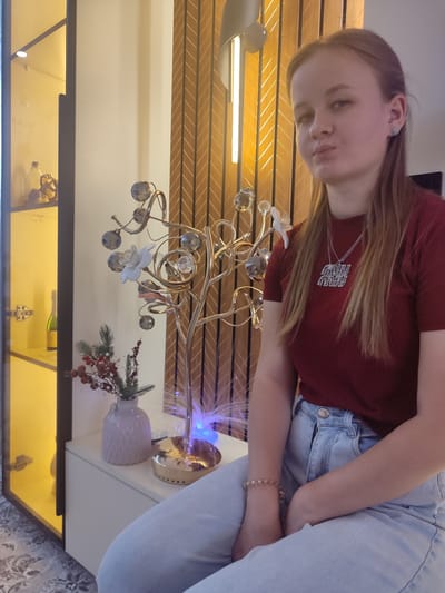 A young woman with blonde hair is sitting on a white cabinet in what appears to be a modern living room. She is wearing a dark red t-shirt and light blue jeans, accessorized with a silver necklace and a beaded bracelet. Her posture suggests she is relaxed and looking towards the camera.

Behind her, a decorative lamp with a golden base and crystal-like embellishments is visible, as well as a vase with a small arrangement of red berries and greenery. To the left, a glass-fronted cabinet with a yellow interior is filled with various decorative items, including bottles. The background features a wall with vertical wooden slats in a light brown hue. The overall lighting suggests an indoor evening setting.

This scene is not located in the city of Chuy, Kyrgyzstan.
