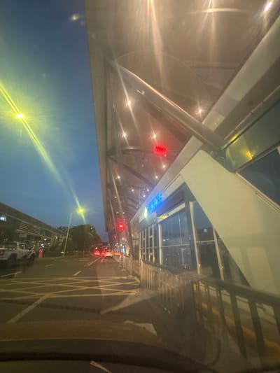 The image depicts an outdoor scene at dusk or night, likely in Xinwu District, China, adjacent to a modern building with a distinctive angled roof structure supported by metal beams. The sky is a deep blue with some light pollution or reflections from streetlights. A bright yellow streetlight with a visible lens flare illuminates the left side of the frame, casting long streaks of light across the sky and the road.
A street runs through the scene, marked with yellow criss-cross markings on the asphalt. Several vehicles are visible on the road, including a white pickup truck on the left, and other cars with illuminated taillights further down the road, indicating traffic. On the right side of the image, the building features large glass windows and an entrance with an automatic sliding door. A blue illuminated sign is affixed above the entrance, displaying text in Chinese characters. Above this, a red digital display is visible, showing numbers and potentially other symbols. A metal railing runs along the walkway in front of the building. The overall environment suggests an urban setting, possibly an transportation hub or a modern commercial area.
