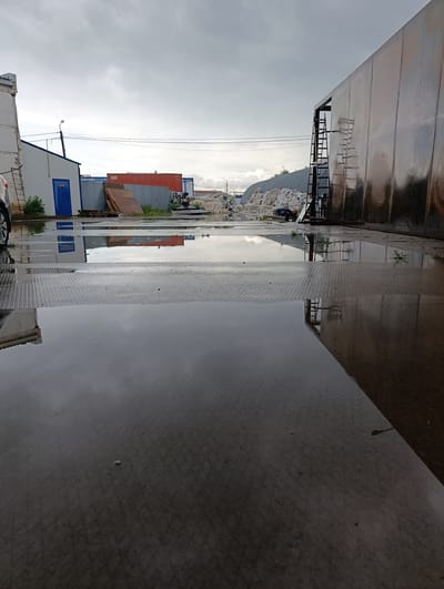 The image depicts an outdoor industrial or storage area. A large, metallic building with a reflective surface occupies the right side of the frame. In the background, there are several smaller structures, including a white building with a blue door and a red shipping container. The ground is covered in puddles of water, suggesting recent rain. A ladder is leaning against the large metallic building. The sky is overcast. There are no people visible in the image. The scene is likely located in a part of Oral, Kazakhstan, that features industrial facilities or storage yards.