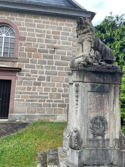 The image shows a war memorial in front of a stone building in Asterode, Germany. The memorial features a stone lion statue seated on a pedestal. The pedestal has an inscription in German that reads: "ZU EHREN DER IM WELTKRIEG 1914-1918 GEFALLENEN HELDEN DIE GEMEINDE ASTRODE" which translates to "In honor of the fallen heroes of the World War 1914-1918, the community of Asterode". Below the inscription, there is a relief of a wreath with an iron cross. To the left of the memorial is the side of a building with a large arched window and a dark door. Green grass and some small yellow flowers are visible in front of the building and the memorial. A dense green tree is present behind the memorial on the right side of the image. The sky is overcast.