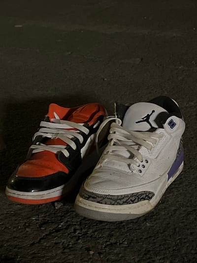 The image shows two sneakers resting on what appears to be asphalt. The top sneaker is a white and gray Air Jordan 3 with purple accents and a black Jumpman logo. The lower sneaker is an orange and black Air Jordan 1. The location is not discernible as being in Lane End, United Kingdom from just the visual data in the image. The lighting suggests it could be outdoors and potentially at night or in dim light.