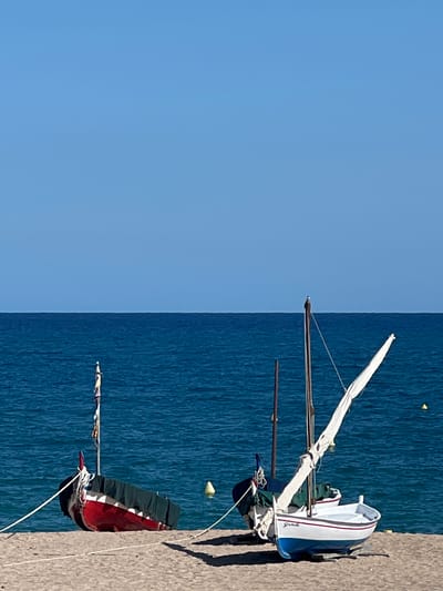 The image displays a tranquil coastal scene under a bright, clear blue sky, characteristic of Sant Pol de Mar, Spain. The composition is horizontally split, with the calm, deep blue sea occupying the left half and a light-colored concrete shore or pier on the right.

Two small sailboats, identified as dinghies, are pulled up onto the shore. The upper boat features a white hull with a distinct blue and narrow red stripe running along its side. Its mast is upright, and a white sail, partially folded or furled, is secured. The word "Grecale" is clearly visible in script on its hull.

Below it, a second boat, smaller and with a vibrant red hull, rests on the shore. Its sail area is covered by a dark green or black canvas. This boat also has a mast, adorned with what appear to be small, colorful pennants or streamers. Both boats are secured by white ropes, some extending into the water or tied to the shore.

The sea exhibits gentle ripples, and two small yellow buoys are visible floating in the water. The bright, even lighting indicates a sunny day, likely mid-morning or afternoon, with no clouds in the sky. No people are present, emphasizing the serene and peaceful atmosphere of the coastal setting.