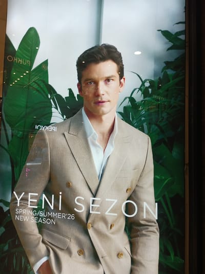 This is a full-frame, eye-level, slightly wide-angle shot of a male model in a light-colored suit, likely a promotional advertisement displayed in a storefront window in Kırıkkale, Turkey. The model is a Caucasian male with short, dark hair, looking directly at the camera. He is wearing a light-colored, double-breasted suit jacket over a white collared shirt. The suit has visible texture, possibly linen or a linen blend, and features two large buttons and lapels.

Behind the model, large green leaves create a lush, tropical backdrop. White text is overlaid on the image. In large, bold font, the words "YENİ SEZON" are prominently displayed, which translates to "NEW SEASON" in Turkish. Below this, in smaller font, are the words "SPRING/SUMMER '26" and "NEW SEASON."

Reflections are visible on the glass surface, indicating it is a display window. These reflections include what appear to be lights and possibly the interior of a shop or the street outside. Some smaller text is also reflected on the glass, with some of it appearing mirrored and partially obscured. One visible mirrored word, likely a brand name, is "IPEKÇI". Another reflected word, also mirrored and partially obscured, appears to be "ARMİNE". The overall impression is a fashion advertisement designed to attract customers to a clothing store. The specific location within Kırıkkale is not discernible from the image itself, but the presence of Turkish text strongly suggests a location in Turkey.