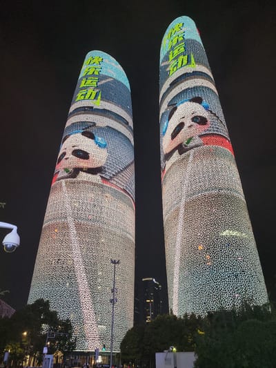 The image shows two illuminated skyscrapers at night in Wuhou District, China. The buildings are covered in a patterned light display with large cartoon images of pandas. The panda on the left building is facing left, and the panda on the right building is facing right. Both pandas have large black eyes, pink cheeks, and are holding green objects in their mouths. The tops of the buildings are adorned with green Chinese characters. The skyscrapers are tall and cylindrical, tapering towards the top. The surrounding environment is dark, indicating it is nighttime. Trees and streetlights are visible at the base of the buildings. A security camera is mounted on a pole to the left of the frame. The light display is vibrant and appears to be a form of architectural projection mapping or LED illumination.