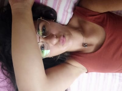 An individual, appearing to be a female, is depicted lying on her back in an indoor setting. Her head is positioned to the left of the frame, with her body extending to the right. She has dark hair and is wearing large, light-colored, cat-eye framed eyeglasses. Her lips are pursed, and she looks directly at the camera. She wears a reddish-brown sleeveless top. A silver-toned necklace with a small, intricate pendant is visible around her neck. Her right arm, from the viewer's perspective, is bent and raised, with the forearm visible in the foreground. She rests on a pillow covered with a white fabric featuring narrow pink stripes. A pink fabric, likely a sheet or blanket, is partially visible beneath her. No identifiable features within the image indicate a specific location in San Marzano sul Sarno, Italy.