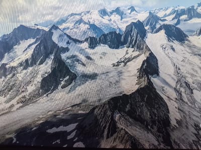 The image shows a vast, mountainous landscape covered in snow and ice. Sharp, jagged peaks rise dramatically, with glaciers flowing down their slopes. The terrain is rugged and appears to be at a high altitude. The sky is partly cloudy, with patches of blue visible. There are no people, structures, or signs of civilization in the image. The scene depicts a remote and wild natural environment. The location in Reftinsky, Russia, is not discernable from the image itself, as the visual information only pertains to a natural mountainous region.