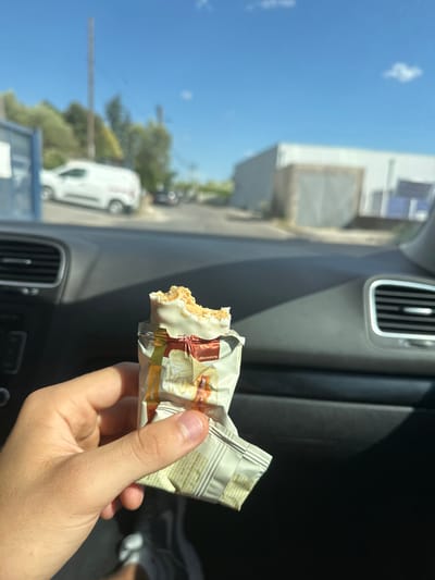 The image shows a close-up of a hand holding a partially eaten white chocolate-covered snack bar with visible nuts and caramel filling. The snack bar is in its packaging, which is crumpled and partially opened. The background, viewed from inside a car, shows a sunny day with a clear blue sky. Trees and a white van are visible in the distance. A building with a grey facade is also in the background. The interior of the car, with its dashboard and air vents, is in the foreground. The location is specified as Alès, France.