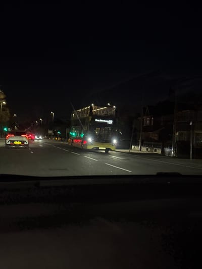 The image captures a nighttime street scene in Bury, United Kingdom, viewed from inside a vehicle looking forward through the windshield. A yellow double-decker bus, operating route 97 between Bury and Shudehill, occupies the right lane, moving towards the camera. Its headlights are illuminated, and green interior lights are visible. Ahead of the camera's vehicle, in the left lane, a white car displays active red taillights. Distant red taillights and white headlights from other vehicles are visible further down the road. The street is a multi-lane road, illuminated by streetlights. To the right of the bus, dimly lit buildings, possibly residential or commercial structures, line the street. The overall environment suggests an active urban or suburban thoroughfare at night. No specific individuals are clearly visible.