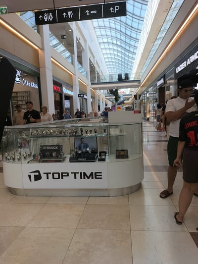 The image captures a scene inside a modern shopping mall, likely in Pardubice, Czechia, given the context of the image provided. In the foreground, a circular glass display case is filled with numerous watches. The brand "G-SHOCK" is prominently displayed on a box within the case, as is another watch brand, "EDIFICE." The display case sits atop a white counter that reads "TOP TIME" in large black lettering, accompanied by a stylized 'T' logo.
Behind the display, a walkway of polished beige tile is visible, leading further into the mall. Several people are present in the scene. A man and woman are seen standing near the display, appearing to be customers. Further back, more shoppers walk through the mall, some carrying bags.
The mall environment is characterized by high ceilings with a distinctive curved, glass-paneled roof, allowing natural light to flood the space. White pillars and the polished floors contribute to a bright and open atmosphere. Retail storefronts line the sides of the mall, with visible signage such as "UNDER ARMOUR" and other unreadable text on various shop facades. Overhead, directional signs indicate "KINO" (cinema) and "P" (parking). The overall impression is one of a bustling commercial hub.