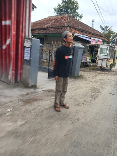The image depicts a male individual standing on a paved surface in Rancamanyar, Indonesia. The individual, appearing to be an adult male, has short grey hair and wears a light-colored skullcap. He is dressed in a black long-sleeved shirt with a red and white YouTube logo on the chest and orange accents on the sleeves, khaki cargo pants, and brown open-toed sandals. His head is tilted upwards and to the right, with his mouth slightly open, indicating an upward gaze.

The immediate foreground consists of a grey, uneven paved surface with areas of darker discoloration, suggesting dirt or moisture. To the left, a tall, vertical structure made of red corrugated metal is partially visible, appearing to be part of a gate or building facade. Adjacent to this and behind the standing individual is a grey concrete pillar, with several white papers affixed to its surface. A black wrought-iron gate is visible behind the individual, leading to a residence.

In the mid-ground, a house with a reddish-brown tiled roof and green fascia is visible. To the right of the house, a small vendor kiosk with a blue awning displaying "MAS CAN...A" and various products is present. A green propane tank and a small wooden table are positioned near the kiosk. Further to the right, a second wheeled vendor cart with a glass display case is observed. Overhead utility wires traverse the upper right portion of the frame. The sky is overcast. A green tree crown is visible above the tiled roof of the house. The overall scene suggests a mixed-use residential and commercial street environment. Motion blur is present across parts of the image, particularly on the ground and the left side.