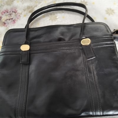 The image shows a black leather handbag with gold-colored hardware. The handbag has two straps and a structured design with visible stitching details. It appears to be sitting on a surface covered with a patterned fabric, possibly a tablecloth or bedspread, featuring floral motifs in pink and light yellow. The background is out of focus, but the overall impression is of an indoor setting. There are no people visible in the image, and no specific activity is taking place. The lighting suggests it is daytime, but no further details about the time of day or weather can be inferred. No visible text is present on the handbag or in the scene. The provided location context of Cupcini, Moldova, does not offer any further clues to the specific setting or content of the image.