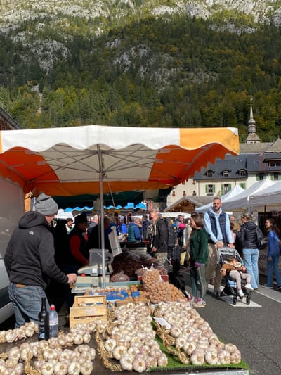 The image displays an active outdoor market in Abondance, France, set against a mountainous backdrop on a sunny day. In the foreground, a market stall under a large orange and white umbrella-style canopy is prominent. Numerous white garlic braids are meticulously displayed on green artificial turf on the stall's counter. To the left, a man wearing a grey beanie, black "The North Face" hoodie, and jeans stands, holding a glass. A black bottle and an Evian water bottle are visible next to a wooden crate behind the garlic. Behind the counter, a vendor in a red vest operates a grey weighing scale, and bags of nuts, including walnuts and chestnuts, are arrayed.
In the mid-ground, a bearded man in a light blue puffer vest pushes a dark stroller containing a young child. Adjacent to him, a woman in a dark green tracksuit walks a black dog on a leash. Numerous other market attendees are seen walking and browsing stalls with white and blue awnings. Buildings with traditional architecture, featuring dark roofs and light-colored facades, are visible behind the market, with a sign partially reading "L'Abbaye." A prominent church spire, indicative of the Abondance Abbey, with a dark, pointed roof and weather vane, rises on the right side of the background. The upper background is dominated by a steep mountain face covered with dense green forest and visible rocky outcroppings.