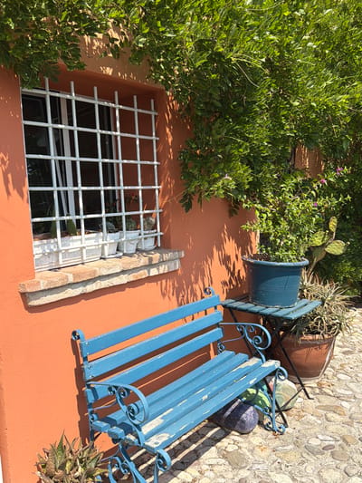 This image depicts a sun-drenched corner of an outdoor seating area, likely in Santarcangelo di Romagna, Italy, given the architectural style and vegetation. An orange-walled building dominates the left side, featuring a window with white bars and a brick windowsill. Several potted plants, including cacti and succulents, are visible on the windowsill, suggesting a Mediterranean climate. In front of the building, a weathered blue wooden bench is positioned on a cobblestone path. To the right of the bench, a small, dark green metal table supports a large blue planter containing a leafy green plant. Behind the table, a terracotta pot holds another plant, possibly a succulent or aloe, and a prickly pear cactus is visible against the wall. Lush green foliage hangs from above, casting dappled shadows on the scene, enhancing the feeling of a peaceful, outdoor retreat.