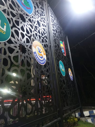 The image shows a black metal fence with an intricate geometric pattern at night. Several circular stickers or emblems are affixed to the fence. From left to right, the visible emblems are: a green oval with the word "LED" in yellow, a blue and white oval with a green leaf and red accents, an orange and blue circular emblem with Indonesian text that appears to read "DIKTIS AINTEK" and "BERDAMPAK", a multicolored oval with abstract shapes, a teal oval with the word "BLU" and "SPEED" in white, and another orange and blue circular emblem similar to the third one. The fence is illuminated by a bright overhead light source, casting a strong glare on the upper right portion of the image. In the background, through the fence, there are blurred lights, including red horizontal streaks that suggest vehicle traffic. The environment appears to be outdoors, possibly near a road or pathway, with some greenery visible at the bottom. The scene is likely in Patrang, Indonesia, given the mention of Indonesian text on the emblems and the context provided.