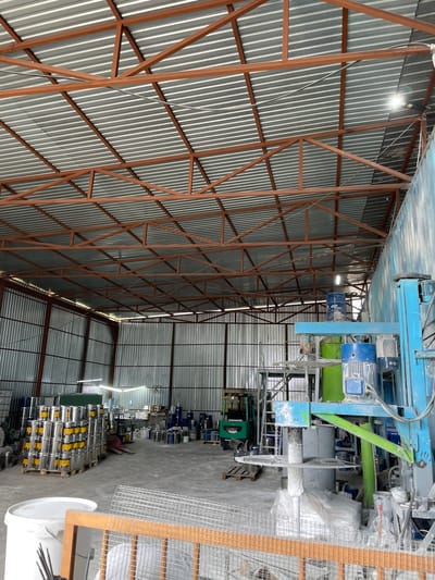 The image shows the interior of a warehouse or industrial facility located in the village of Kenen Azirbayev, Kazakhstan. The structure has a corrugated metal roof supported by a reddish-brown metal frame. The walls are also made of corrugated metal. In the foreground, a large white bucket is visible, along with a wooden frame and wire mesh. To the left, stacks of metal cans with yellow labels are organized on wooden pallets. In the center and right of the image, there is a blue industrial mixing machine with various components, including motors and tanks, one of which is green. A small green forklift is parked in the middle ground. Various other containers and equipment are scattered throughout the warehouse. The lighting appears to be from overhead fixtures. There are no people visibly present in the scene.