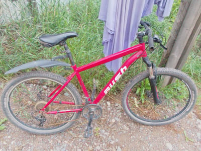 A red bicycle with a black seat is leaning against a wooden fence post, partially obscured by tall green grass and a folded purple fabric. The bicycle has a visible "RUSH" logo on its frame. The front wheel is turned slightly to the right. The ground is covered with small stones and dirt. The scene appears to be outdoors, possibly in a rural or overgrown area.