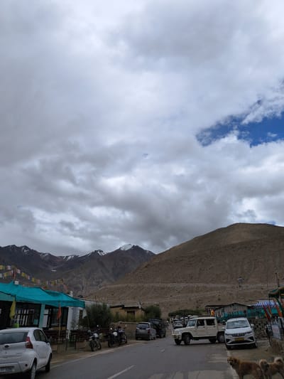 A street scene in Khardung, India, features a road with several vehicles, including a white SUV, a white utility vehicle, motorcycles, and a white car. Mountains with snow-capped peaks form the backdrop, under a cloudy sky with patches of blue. Buildings line the side of the road, some appearing to be shops or restaurants with colorful awnings. Prayer flags are strung across some of the buildings. Two dogs are visible in the foreground on the right side of the image. The overall impression is of a roadside settlement in a mountainous region.