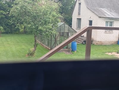 The image depicts a rural backyard scene in Maļutki, Latvia. In the background, a light-colored brick house with a grey corrugated roof is visible. To the left of the house, a greenhouse with a metal frame and translucent panels is situated on a patch of grass. Adjacent to the greenhouse is a large blue plastic barrel. In the foreground, a thick, dark horizontal bar obstructs the view, obscuring the lower portion of the scene. A wooden or metal structure, possibly a ramp or railing, extends diagonally from the bottom right towards the center of the image. The yard is covered in green grass, with scattered trees and foliage. The overall impression is one of a modestly maintained outdoor space at a private residence.