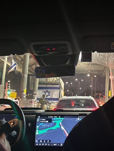 The image shows the interior of a vehicle at what appears to be a checkpoint or border crossing. Outside the vehicle, an Israeli flag is visible, and a person in military uniform holding a rifle is standing. There are other vehicles in the background, as well as traffic lights and signage, some of which appears to be in Hebrew. The dashboard of the vehicle shows a navigation screen with a map and a temperature reading of 24.5 degrees Celsius. The environment suggests a nighttime or dusk setting, with artificial lighting illuminating the scene. The overall context points to a location within the Israeli settlement of Modi'in Illit in the Palestinian Territories.