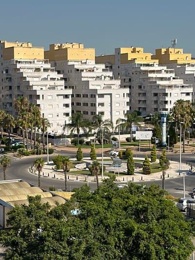 This image captures an aerial view of a modern urban landscape, featuring a roundabout at the intersection of several roads. Dominating the scene are large, multi-story apartment buildings with a distinctive architectural style, characterized by a lighter colored base and bright yellow upper sections. The buildings have numerous balconies and windows, indicating residential occupancy. The roundabout itself is landscaped with manicured bushes, palm trees, and conical shaped trees, surrounding a central fountain. Several cars are visible on the roads, indicating traffic flow. Streetlights are positioned along the roadside. In the background, the sky is clear and blue, suggesting a sunny day. The scene appears to be located in a well-developed area with contemporary infrastructure and landscaping.