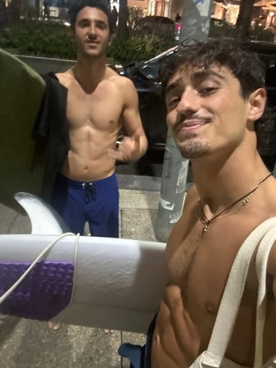 Two shirtless young adult males are depicted at night in an urban-coastal environment, specifically Bondi Beach, Australia. The individual in the immediate foreground on the right, with dark curly hair, a mustache, and a goatee, smiles directly at the camera. He wears two thin necklaces and has a light-colored canvas bag over his left shoulder, holding a white surfboard partially visible in the bottom right, which features a purple traction pad and a white fin. The second individual, positioned in the mid-ground on the left, also with dark curly hair, looks towards the camera with a neutral expression while making a hand gesture. He wears blue swim shorts and has a dark fabric draped over his left shoulder, holding a green surfboard under his left arm. The background shows a dark vehicle, illuminated streetlights, and distant building lights, indicating a nighttime setting. The ground beneath the individuals is paved.