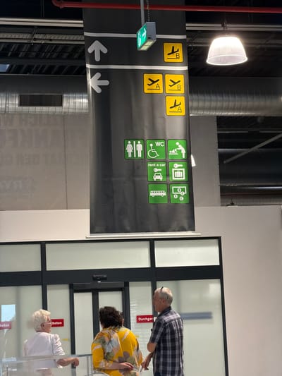The image depicts an indoor scene, likely an airport in Lautzenhausen, Germany, based on the presence of a directional sign and people. The sign, which hangs from the ceiling, provides guidance to Terminals A and B, restrooms (WC), access for individuals with disabilities, car rentals, and public transportation (indicated by a bus icon). The sign also features directional arrows pointing left and downward.
Three individuals are visible in the background. One man in a plaid shirt leans towards a service window. Below him, a person wearing a yellow patterned shirt is facing towards the window as well. Next to them an elderly person in a white shirt is looking towards the window too. The window frame is black with white panes.
The environment includes industrial elements such as exposed ventilation ducts, supporting the airport or transportation hub context. The overall lighting is bright and industrial.
