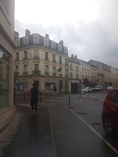This is a photograph taken from a street-level perspective, looking up at buildings and the sky. The image is oriented vertically. In the foreground, a person in dark clothing is walking on a wet street, possibly after rain, towards the right of the frame. Their feet are visible, one in a white sneaker. The street appears to be a city street with buildings lining one side.
The buildings are multi-story, with architectural styles suggesting an older European city. The facades are light-colored, with many windows, some featuring shutters. Some of the buildings have balconies with ornate railings. There are shops visible at the ground level, with signs. One sign clearly reads "PHARMACIE" in large blue letters, and another advertisement above it shows a woman's face with makeup. Another sign above the pharmacy reads "CAUDALIE".
Further up the street, more buildings are visible, also appearing to be residential or commercial. Cars are present on the street; a red car is partially visible in the upper right corner, and other cars, including a white van and a white car, are further down the street. A pedestrian crossing is marked on the asphalt.
The sky is overcast with a bright, diffused light, indicating it is daytime, likely late morning or afternoon, with cloudy weather. The wetness of the street suggests recent rain. The overall impression is that of a typical urban scene in a European city. There are no strong emotional cues conveyed by the image.