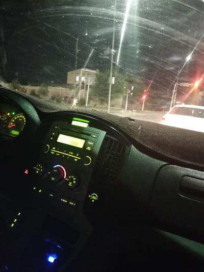 The image is taken from inside a vehicle at night, looking out through the windshield. The dashboard is visible in the foreground, showing a car radio display that reads "FM 99.2", a speedometer, and various buttons and controls for the car's climate system and other functions. The windshield is dirty with streaks and dust, reflecting the lights of the city. Outside, streetlights and traffic signals are visible, as well as the outlines of buildings and trees. A white car is seen in front of the vehicle. The setting appears to be a road in Turkistan, Kazakhstan, given the visible streetlights and architectural style of the buildings. There are no people visible in the image. The overall impression is of a typical urban night scene from within a moving vehicle.
