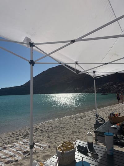 The image shows a beach scene in Unknown, Mexico, viewed from under a white canopy tent. The turquoise water reflects the sun, and a dark, rocky mountain rises in the background. On the right side of the frame, a black folding table holds items including cups, a basket, and a radio. A striped towel lies on the sand. There are people in the distance.