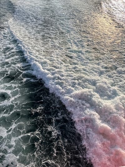 The image displays an overhead view of turbulent sea water in Palaiopoli, Greece. A prominent, diagonal line of dense white foam and breaking wave activity traverses the frame from the lower left to the upper right. To the left of this foam line, the water exhibits a dark blue-green coloration with visible, swirling wave patterns and dispersed patches of lighter foam. To the right of the primary foam line, the water is extensively agitated and appears as a mass of white, churning foam. Sunlight reflects off the water in the upper right portion, creating a bright, shimmering effect. A subtle pinkish coloration is present within the foam in the lower right quadrant. No individuals, vessels, or other identifiable man-made objects are visible within the depicted environment.