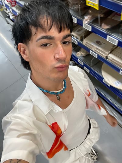 A medium close-up selfie captures a young individual, likely male, with dark, slightly wet hair, looking directly at the camera with a neutral expression. The image is rotated 90 degrees clockwise. The person's skin has a dewy appearance. They are dressed in a white ribbed tank top, worn beneath an open white long-sleeved shirt featuring abstract orange and red patterns on the sleeves. A distinctive necklace adorns their neck, composed of blue and white stone beads and a small silver charm resembling an animal or symbol.
The background suggests an indoor commercial or storage environment, possibly a store or warehouse. Visible are blue metal shelving units, laden with various items, some enclosed in clear plastic packaging. A light beige roll of material and a yellow-packaged item are discernable on different shelves. The floor is light grey or white, and the area is brightly illuminated by artificial lighting.
Additional details include a partially visible tattoo on the person's left forearm, comprising abstract lines. The lower body shows white pants, likely joggers, and one foot wearing a sneaker with a prominent black and white pattern, possibly animal print. No readable text is clearly visible within the frame. The location context for this media file is Su Masu/Elmas, Italy.