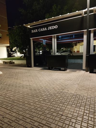 The image captures a nighttime view of the exterior of "BAR CASA JEDO" in Motril, Spain. The bar features a black facade with white lettering spelling out its name. A white awning extends over the entrance, supported by white pillars. To the left of the bar, a white van is parked on a grassy area next to a concrete bench. In front of the bar, on a paved area made of interlocking square tiles, are several black wheeled containers. The background reveals trees and buildings, illuminated by artificial light. The overall scene suggests a commercial establishment in an urban setting at night.