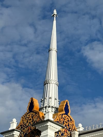 The image shows a detailed view of the spire of the Ostankino Tower in Moscow, Russia. The spire is tall and slender, with a fluted design and decorative elements near its base. Below the spire, the ornate roof of the tower is visible, featuring intricate golden and blue tilework, as well as architectural embellishments. The sky above is a vibrant blue, dotted with fluffy white clouds. The perspective is from below, looking up towards the top of the tower.