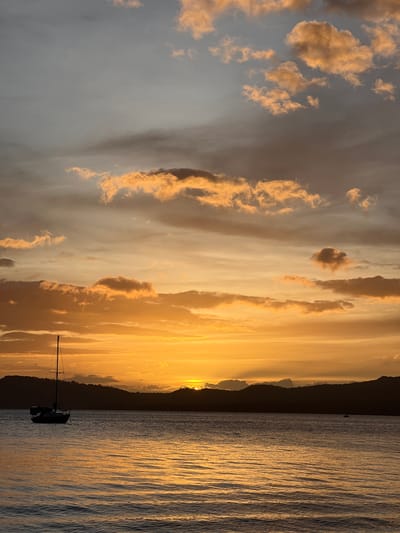 A sailboat is moored in a body of water with a dark, mountainous horizon silhouetted against a dramatic sunset. The sky is filled with clouds that are illuminated by the setting sun, creating a gradient of orange, yellow, and gray hues. The water reflects the colors of the sky, creating a shimmering effect. The scene appears to be a tranquil evening in Altagracia, Nicaragua, as the sun dips below the horizon, casting long shadows and bathing the landscape in warm light.