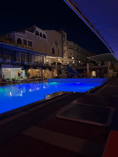 The image depicts a nighttime scene at a resort or hotel in Ayvalık, Turkey, featuring a brightly lit swimming pool. The pool is illuminated with vibrant blue underwater lights, reflecting on the water's surface. Several lounge chairs are arranged around the pool area, some appearing unoccupied and others perhaps with people out of frame.

The background shows multi-story buildings with balconies and windows, suggesting hotel accommodations or apartments. One building exhibits architectural details reminiscent of traditional Turkish design, with arched window openings and decorative elements. Another building appears more modern, with a stacked balcony structure. A blue fabric canopy extends from the top of the image, casting a shadow and adding to the ambient lighting.

A waterslide, possibly for children, is visible near the edge of the pool, adding a playful element to the scene. The ground surrounding the pool is paved with tiles in varying shades of brown and dark gray. A square manhole cover is embedded in the paving stones in the lower right portion of the image. The overall atmosphere is serene and inviting, illuminated by artificial light against the dark night sky. No people are clearly visible within the frame.