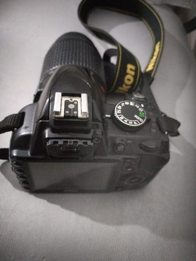 A top-down, slightly angled shot of a black Nikon DSLR camera is placed on a textured, light-colored surface. The camera strap, featuring the Nikon logo in yellow and black, is draped over it. The camera's mode dial is visible and set to "GUIDE" mode. Also visible are the hot shoe mount, a flash connector, and part of the LCD screen and body. The background is blurred and appears to be a soft surface. The lighting is somewhat dim, creating soft shadows. There are no people or other objects clearly discernible in the scene. The location in Moara Domnească, Romania is not indicated by any visible elements within the image.