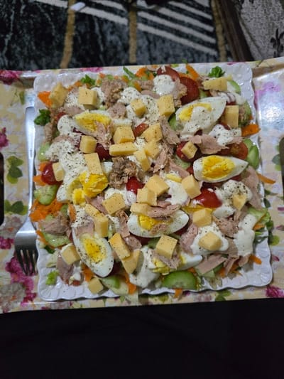 A square white plate holds a tuna salad. Halved hard-boiled eggs, cheese cubes, chopped tomatoes, and cucumber slices are mixed with tuna and a creamy sauce. Shredded carrots add a touch of orange. Black pepper is sprinkled on top. The plate sits on a floral-patterned tray, and a fork rests on the lower left corner.