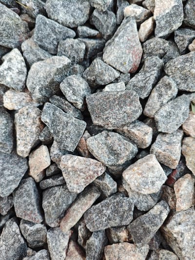 The image displays a close-up, top-down view of a pile of grey, angular gravel. The stones vary in size and shape, with some appearing larger and more irregular than others. Their surfaces are textured and speckled with flecks of black, white, and subtle hints of reddish-brown. The lighting suggests it is daytime, with no indication of specific weather conditions. The setting appears to be outdoors, possibly a pathway or construction area, given the nature of the material. No people, text, or discernible events are visible. The overall impression is one of a natural, rough texture. The location context is Краснадворскі сельскі Савет, Belarus.