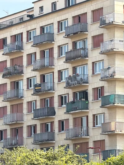 The image depicts the facade of a multi-story building, likely an apartment complex, in Antony, France. The architecture features repeating balconies with metal railings and reddish-brown shutters, interspersed with windows. The building's exterior is clad in a light-colored material, possibly stone or stucco, with a grid-like pattern of darker lines suggesting structural divisions or decorative elements.

A prominent feature is a black and yellow real estate sign hanging from one of the balconies. The visible text on the sign reads "21 CENTURY" followed by "À VENDRE" and a phone number, indicating that an apartment is for sale. The sign is positioned on the balcony of a unit with a metal grate and what appears to be a window.

The image is taken from a slightly angled perspective, looking upwards at the building. The lighting suggests it is daytime, with sunlight casting shadows and highlighting the textures of the building's facade. To the right of the building, lush green foliage from a tree or shrub encroaches on the frame, adding a natural element to the urban scene. The overall impression is one of residential architecture under a clear sky, with a commercial aspect introduced by the "for sale" sign. No people are visible in the image.
