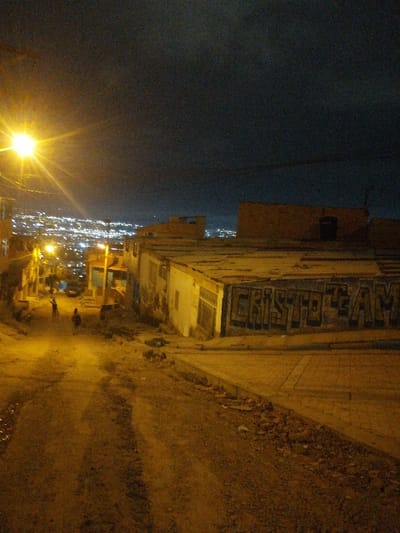A dimly lit, downward sloping dirt road at night. A bright streetlamp casts a yellow glow on the scene, illuminating figures in the distance. Houses flank the road on either side, some with graffiti reading "CRISTO TEAM." In the background, a city lights up the horizon under a dark sky. The scene evokes a sense of urban poverty and quiet isolation.
