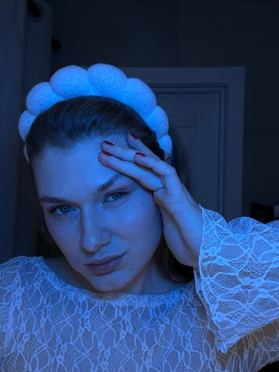 A woman with blue eyes and red nail polish lies sideways, her hand resting on her forehead. She wears a lace-patterned garment and a white, spherical headband. The lighting is dim, casting a blue hue. A portion of a doorframe is visible in the background, slightly out of focus. Part of a surface is present on the left.