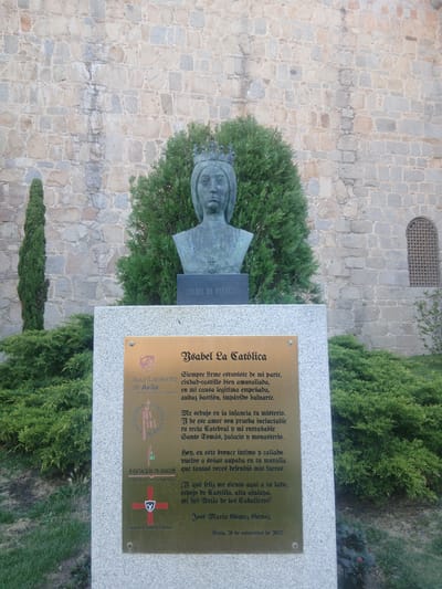 The image shows a bronze bust of Isabella I of Castile, located in Ávila, Spain. The bust is mounted on a stone pedestal with a plaque below it. The plaque is made of bronze and contains text in Spanish. The text mentions "Ayuntamiento de Ávila," indicating the city council of Ávila, and references Isabella the Catholic. Behind the bust and pedestal is a stone wall, characteristic of Ávila's medieval architecture, and some greenery, including trees and bushes. The overall scene appears to be an outdoor monument or memorial.