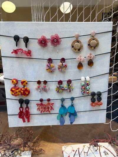 This is a close-up, eye-level photograph of a display of assorted earrings in a shop window in Würzburg, Germany. The earrings are arranged on a white board suspended by black cords. The board is approximately 1 meter tall and 1 meter wide. The earrings are diverse in style, color, and material, including floral designs, straw hats, beaded circles, mosaic patterns, clear and opaque plastic shapes, and coral-like structures. Some earrings feature natural materials, while others are made of plastic or glass beads. The backdrop behind the display board consists of a white macrame net, suggesting an outdoor or artisanal market setting. Below the display, a textured brown fabric or burlap covers a surface, and some dried branches or driftwood are placed in the foreground, adding a natural or nautical theme. The lighting suggests it is daytime, possibly with some artificial light contributing to the scene. No people are visible in the frame.